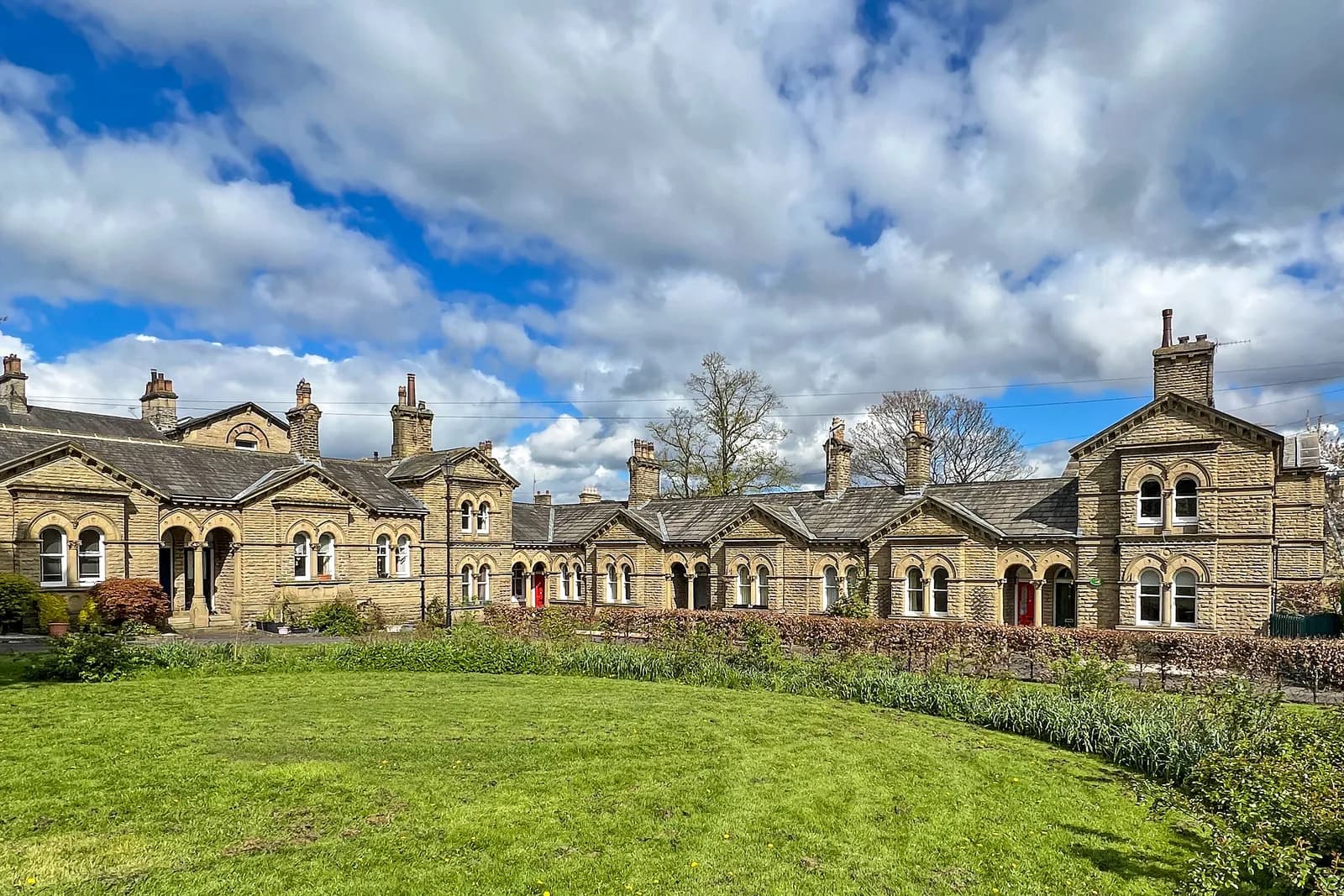 A long, ornate stone building with arched windows and red doors sits behind a vibrant green lawn under a partly cloudy sky.
