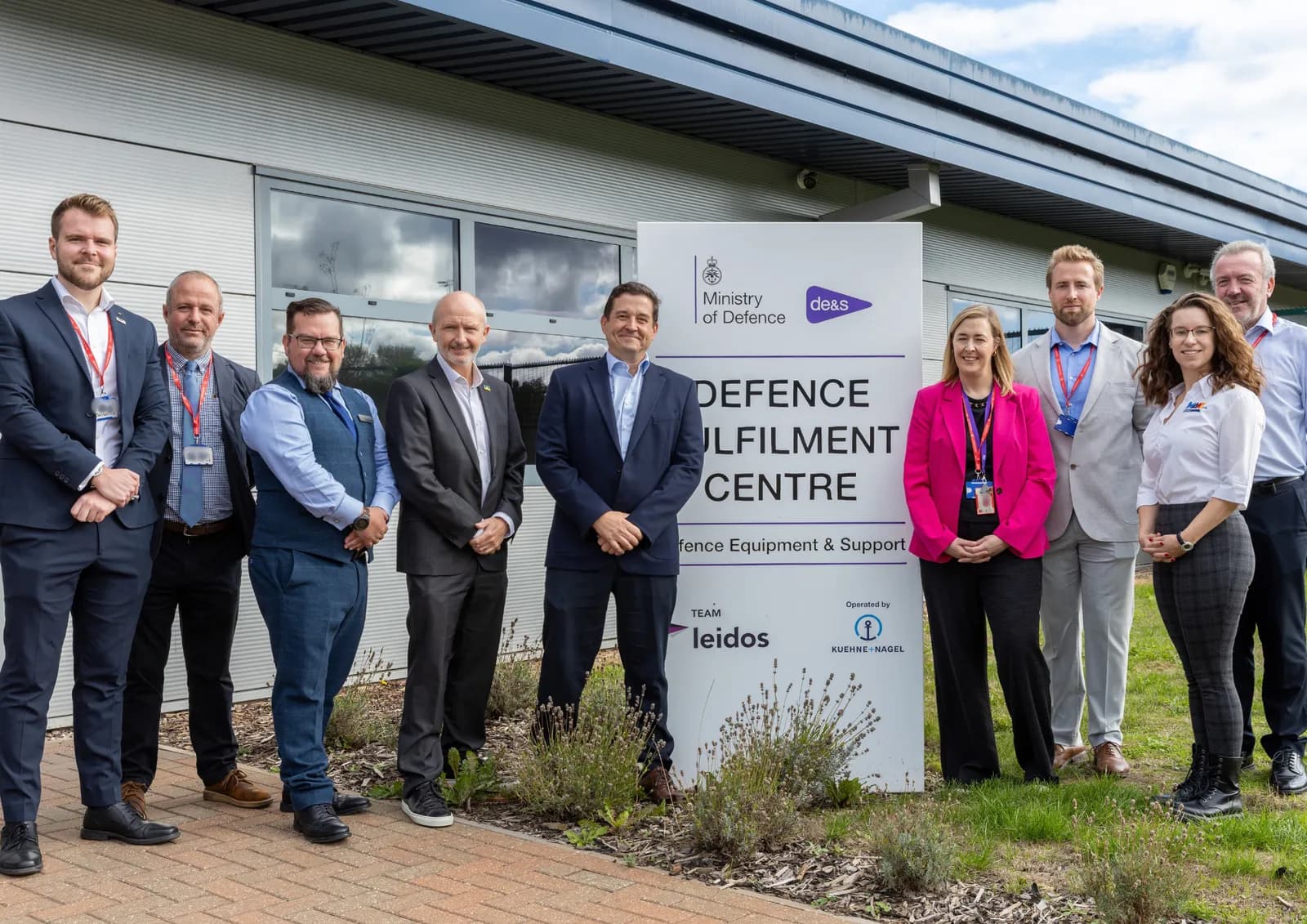 A group of six men and three women in business attire pose for a photo in front of a building and a sign for the Ministry of Defence Defence Fulfilment Centre.