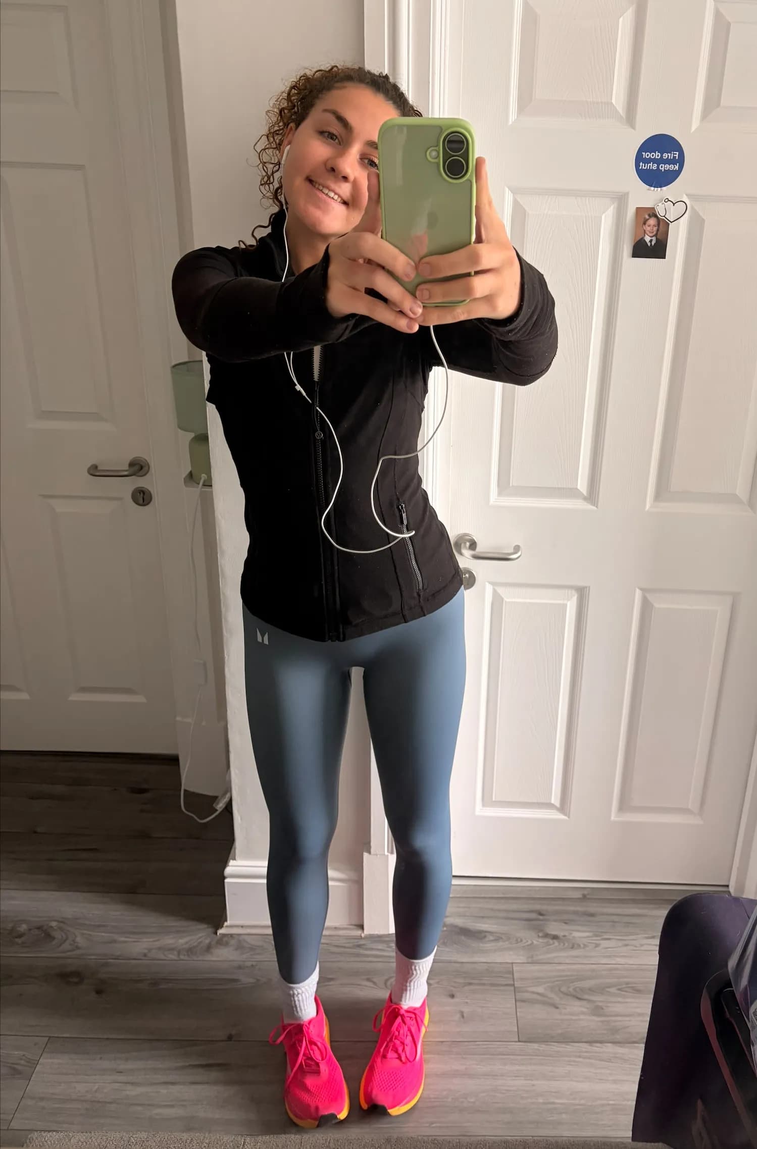 A young woman with curly brown hair, wearing a black athletic jacket and blue leggings with bright pink running shoes, smiles while holding a light green phone to take a mirror selfie.