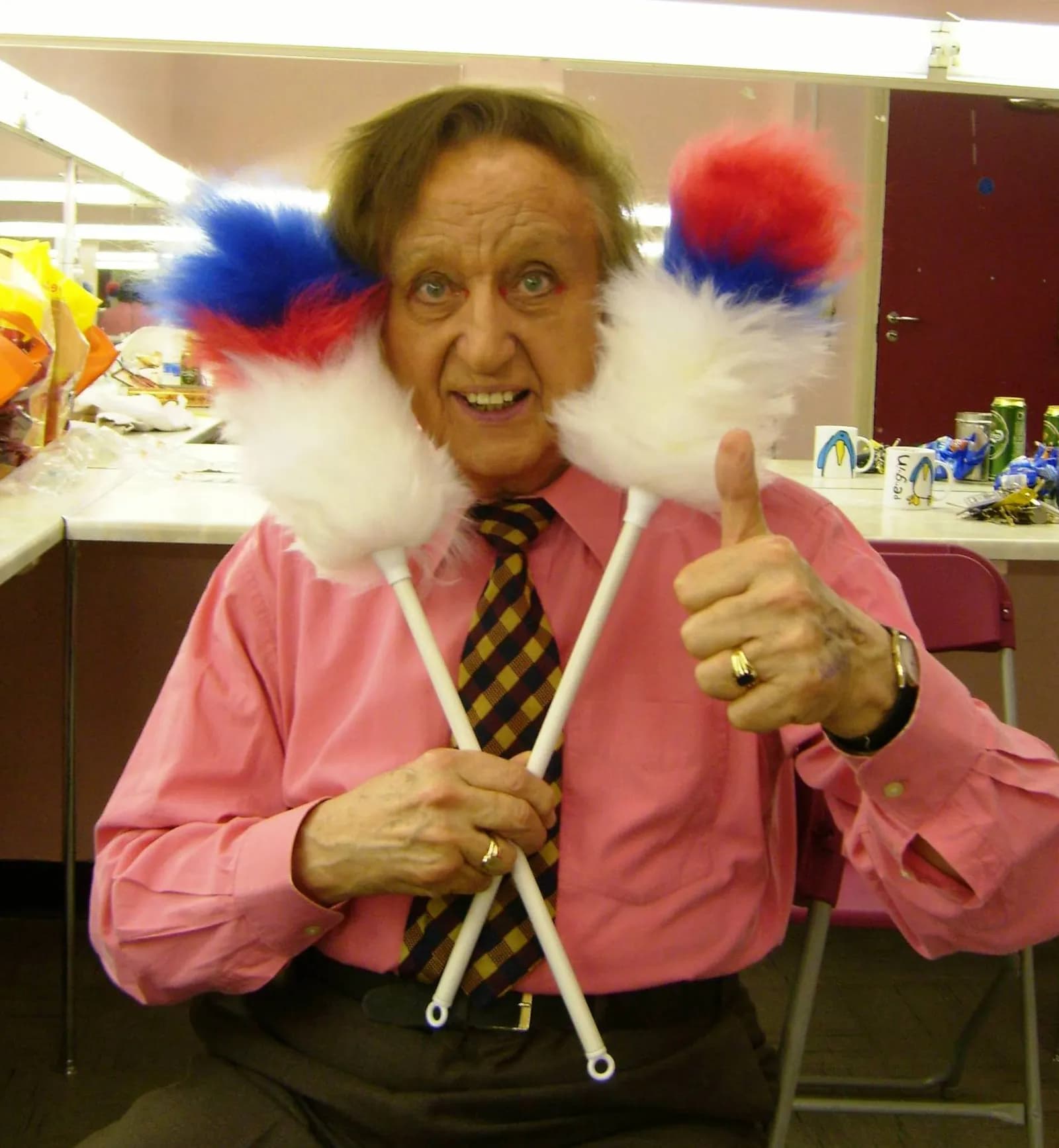 A smiling man with tousled hair, wearing a pink shirt and striped tie, holds two feather duster sticks with red, white, and blue fluff while giving a thumbs up.