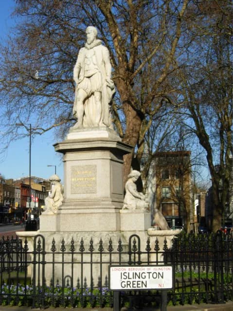 A statue of a man in historical attire stands tall on a stone pedestal with smaller statues at its base, surrounded by trees with bare branches against a clear blue sky, with a street view in the background.