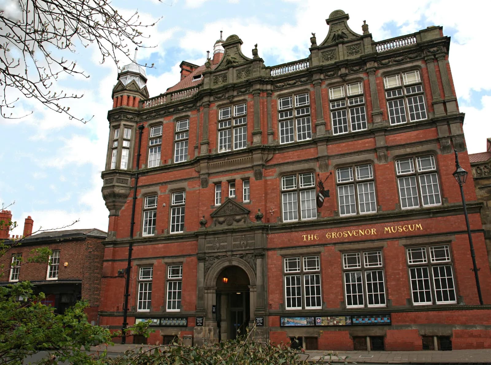 The Grosvenor Museum building, an ornate red brick structure with many white-framed windows and a prominent entrance arch, stands under a cloudy sky.