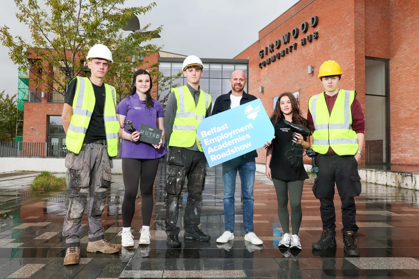 Six people, some wearing safety vests and hard hats, stand in front of a brick building, holding signs related to the Belfast Employment Academies Roadshow.
