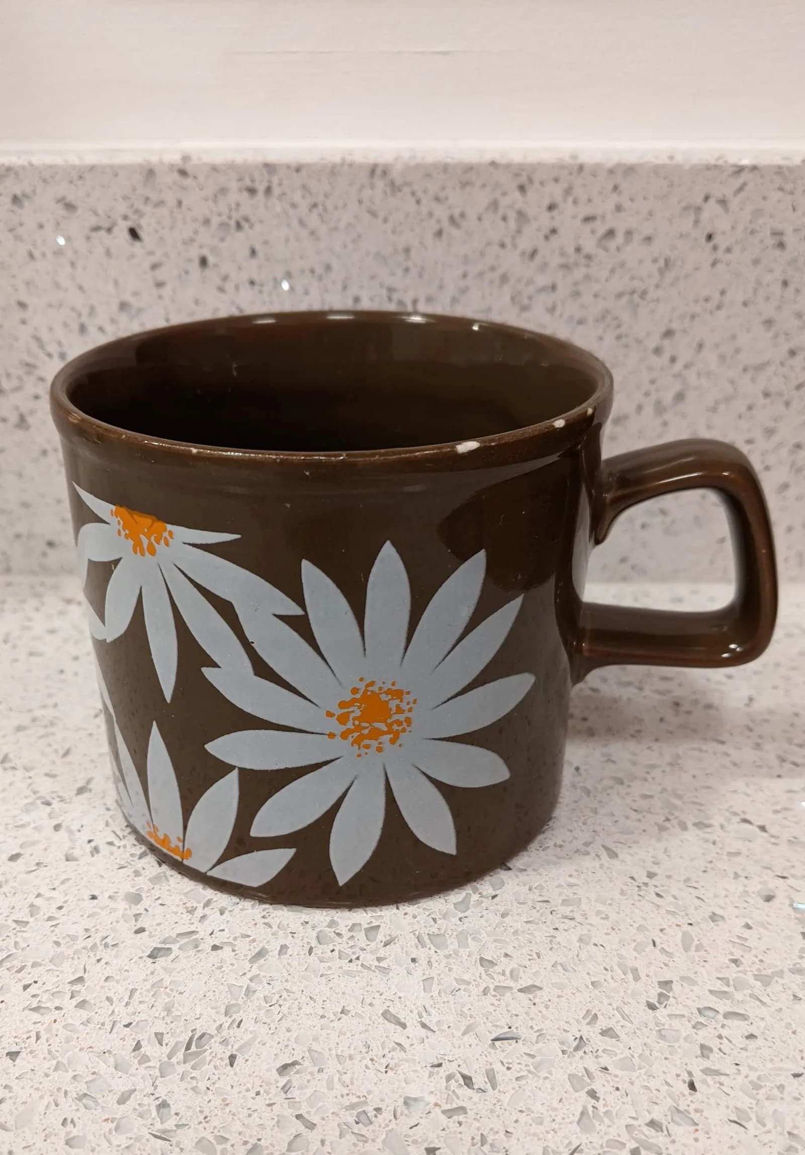 A brown ceramic mug with light blue and orange daisy designs, sitting on a speckled white countertop.