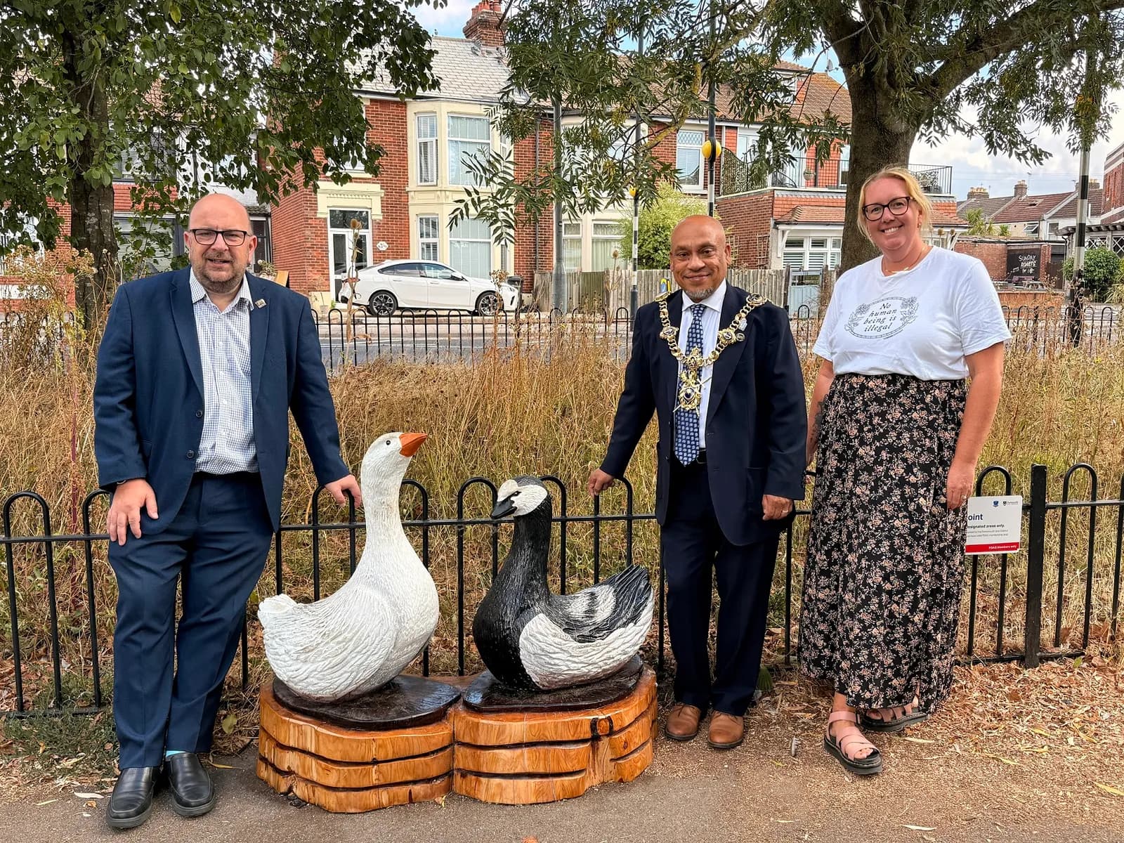 Three people stand outdoors near two goose sculptures on wooden bases behind a black fence, with buildings and a street in the background.