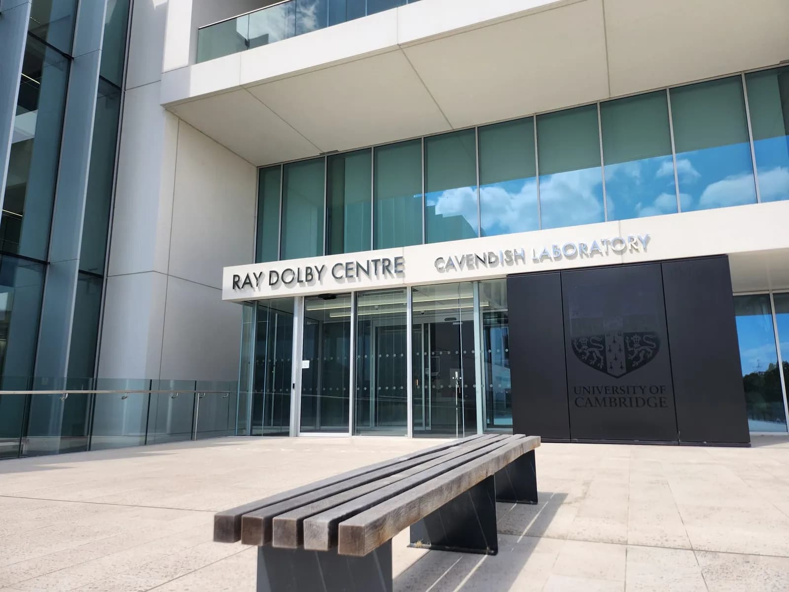 A modern building entrance with "RAY DOLBY CENTRE CAVENDISH LABORATORY" above the doorway and the University of Cambridge logo on a dark panel to the right, with a wooden bench in the foreground.