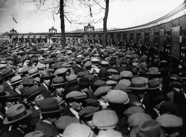 A large crowd of men wearing hats are gathered outdoors, with a curved building or structure and bare trees in the background.