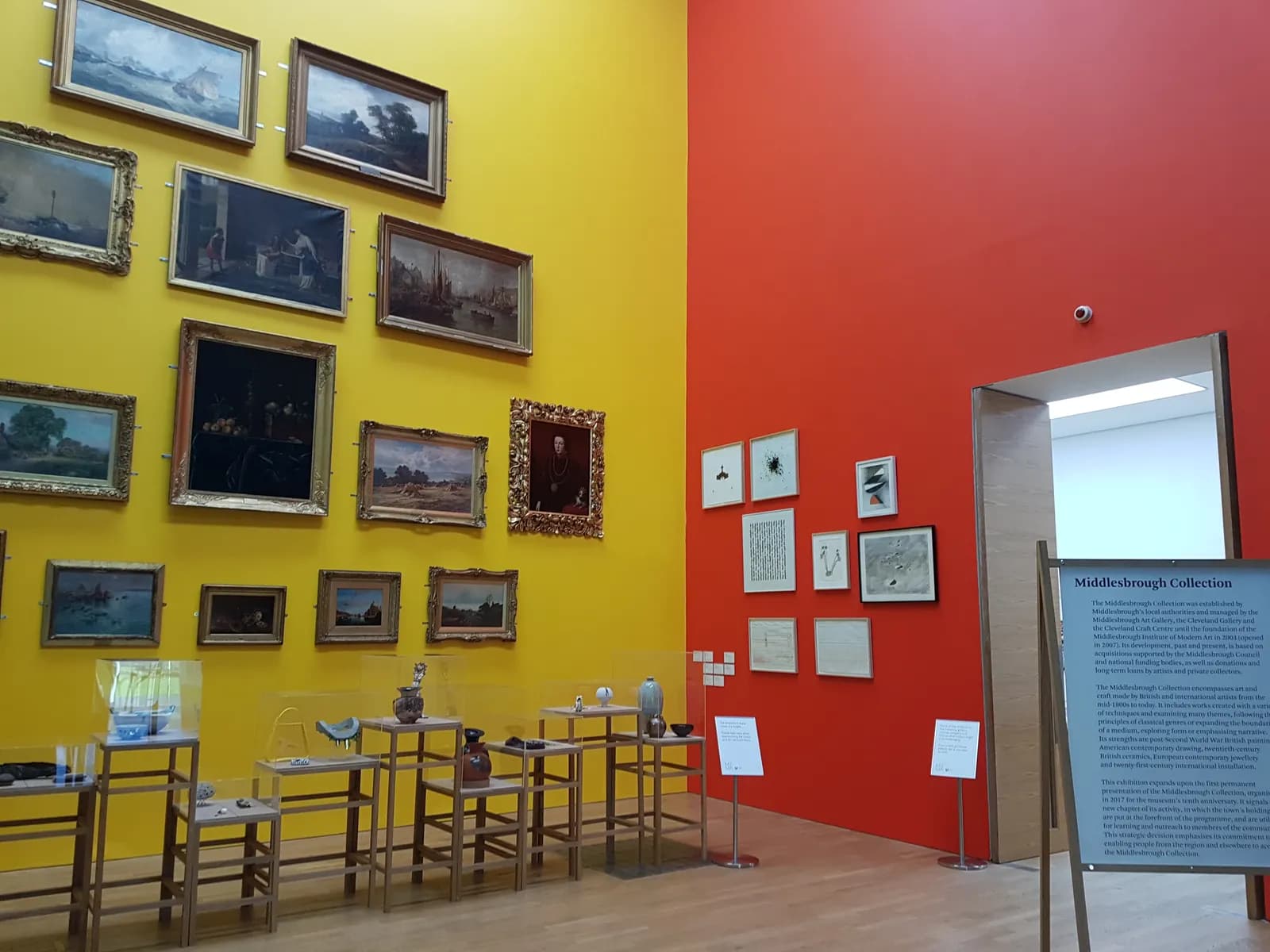 A gallery space with a bright yellow wall on the left adorned with numerous framed paintings, and a vibrant red wall on the right featuring a collection of smaller framed art pieces and an open doorway, with display cases showcasing sculptures and artifacts in the foreground.