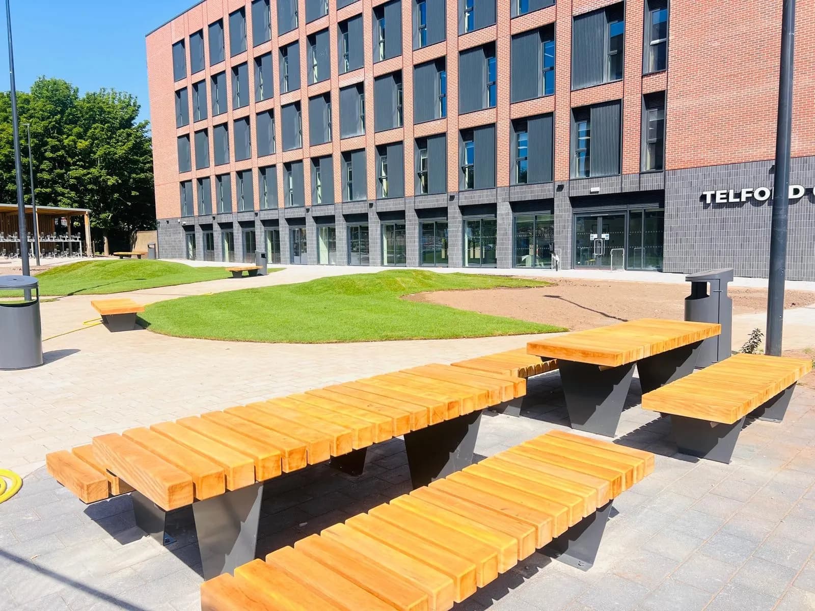 A modern building with large windows and a brick facade stands behind an outdoor seating area featuring light wooden picnic tables and benches on a paved patio with small grassy mounds.