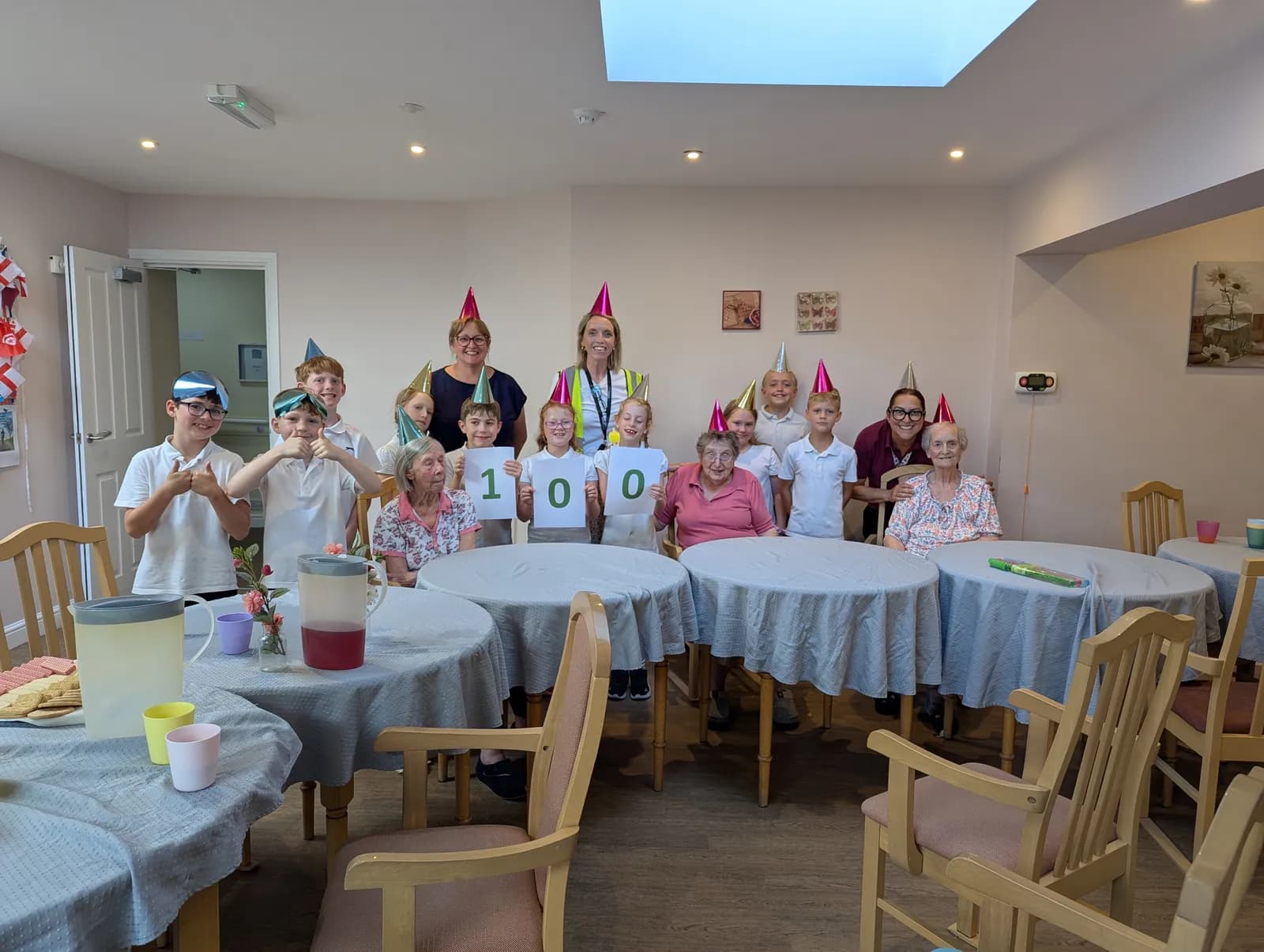 A group of adults and children wearing party hats pose for a photo around a table, with some children holding signs that spell out "100".