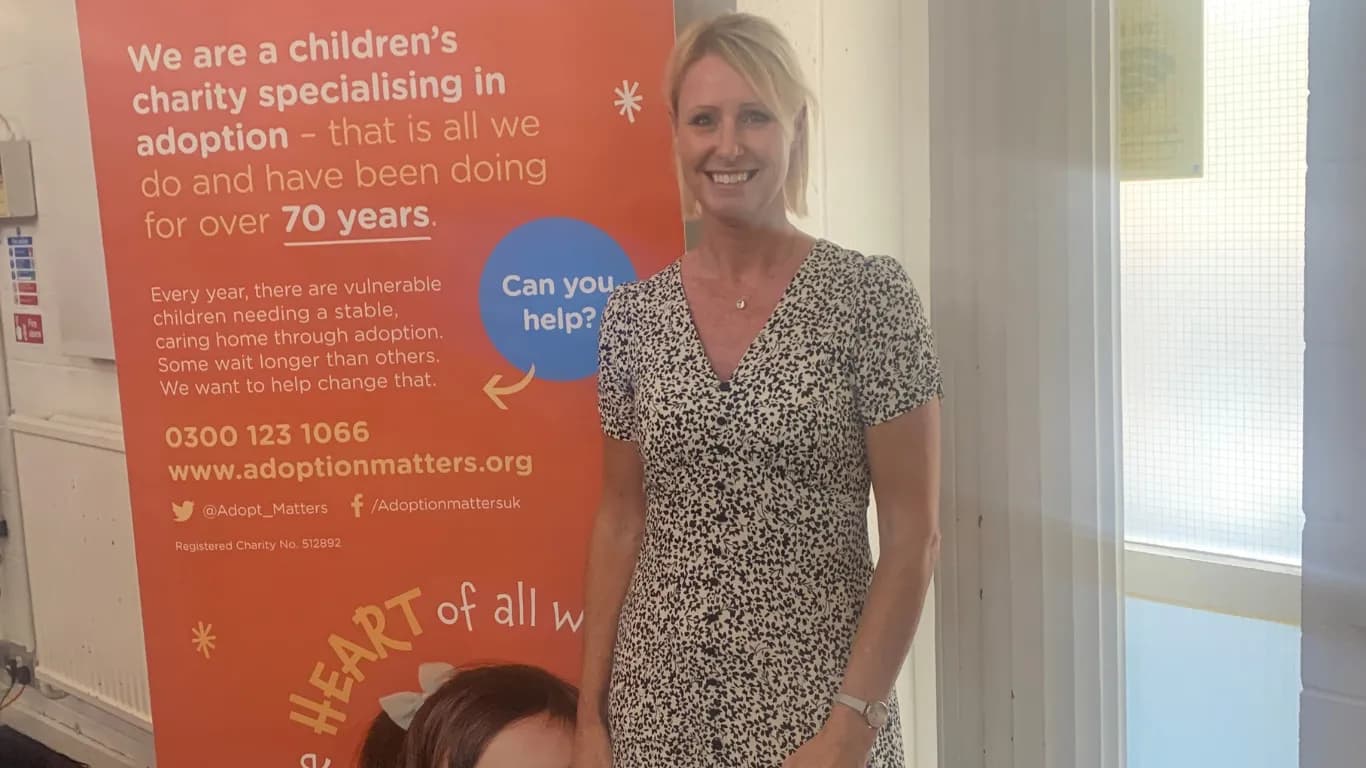 A smiling blonde woman wearing a polka dot dress stands next to an orange banner advertising an adoption charity.