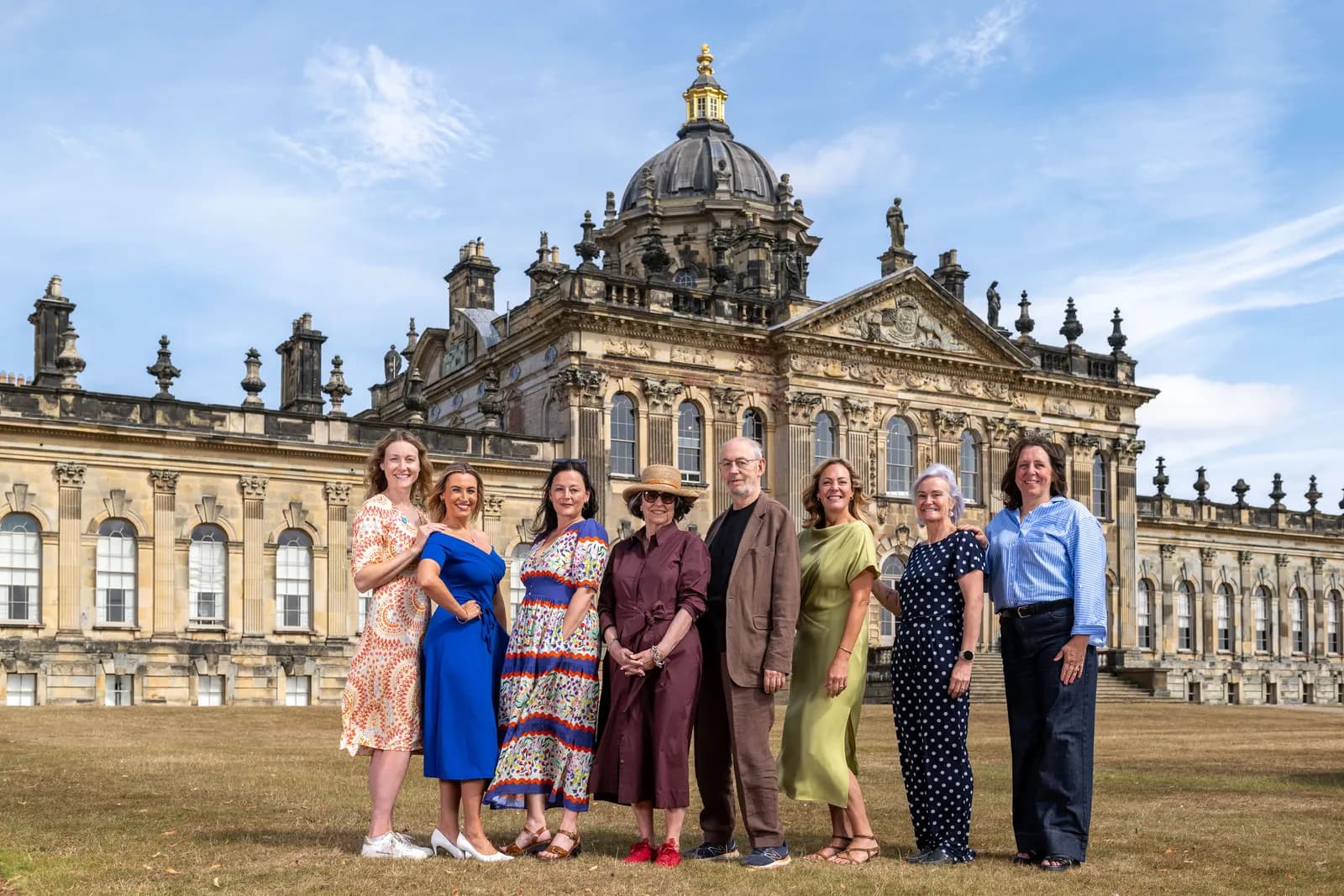 A group of seven women and one man stand on a grassy lawn in front of a grand, ornate Baroque-style building with a large dome.