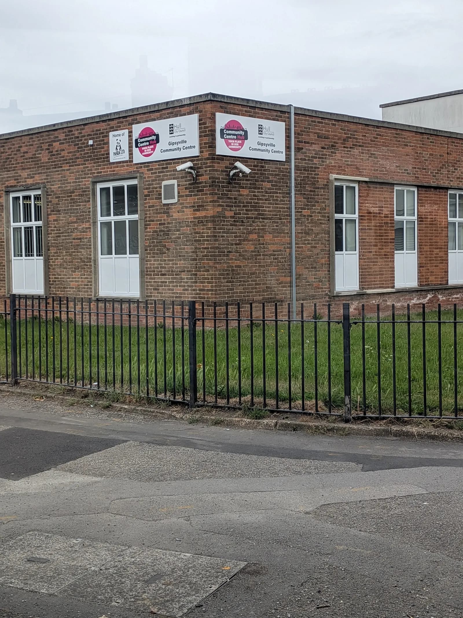 A brick building with white framed windows and two prominent signs for the 'Gipsyville Community Centre' and 'Community Centre Hub,' viewed from across a road with a black metal fence separating it from a patch of grassy ground.