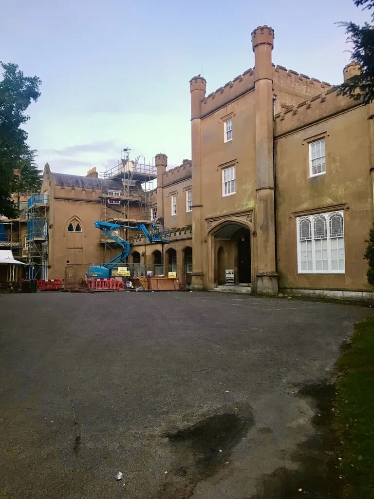 A large, old, tan-colored building with multiple windows and turret-like structures on its roof, partially covered in scaffolding and machinery, sits behind an empty asphalt lot.