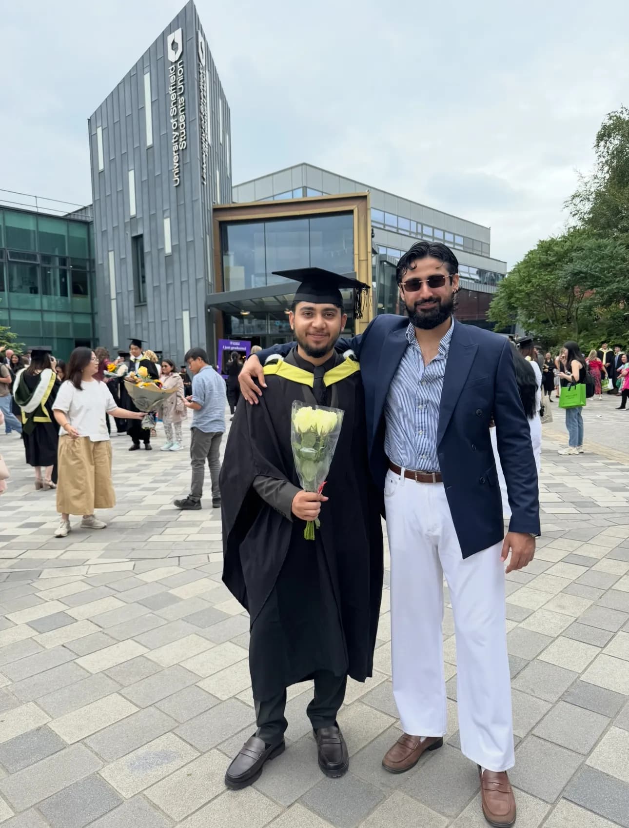 A graduate in a cap and gown holding white roses stands next to a man in a suit, both smiling at the camera, with a modern university building in the background.