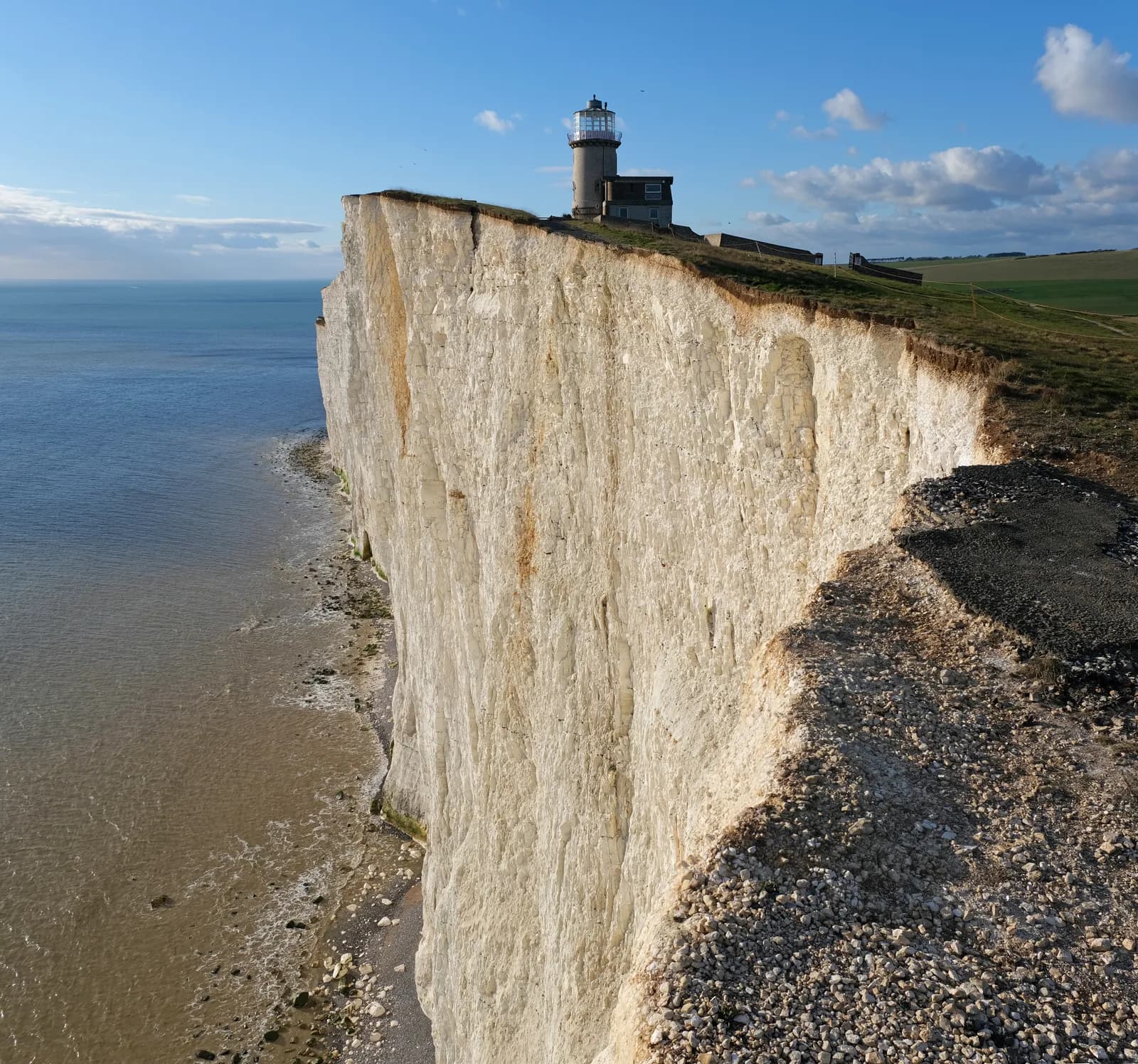 A lighthouse and building are perched on a tall white cliff overlooking a deep blue sea under a bright sky with scattered clouds, with green fields stretching into the distance on the right.