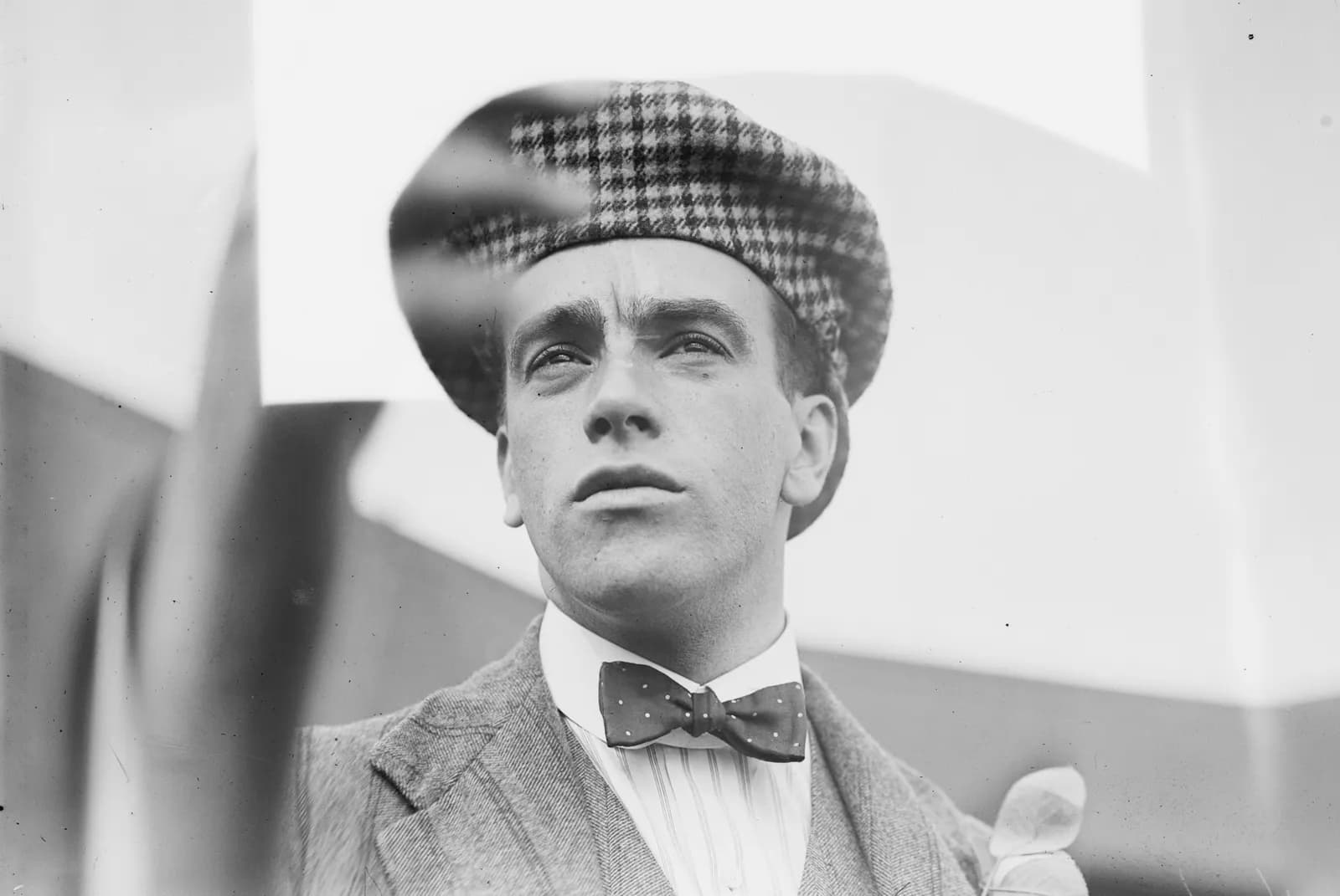 A man in a plaid flat cap, suit jacket, and bow tie looks upwards, with part of his head obscured by a reflection or glare.