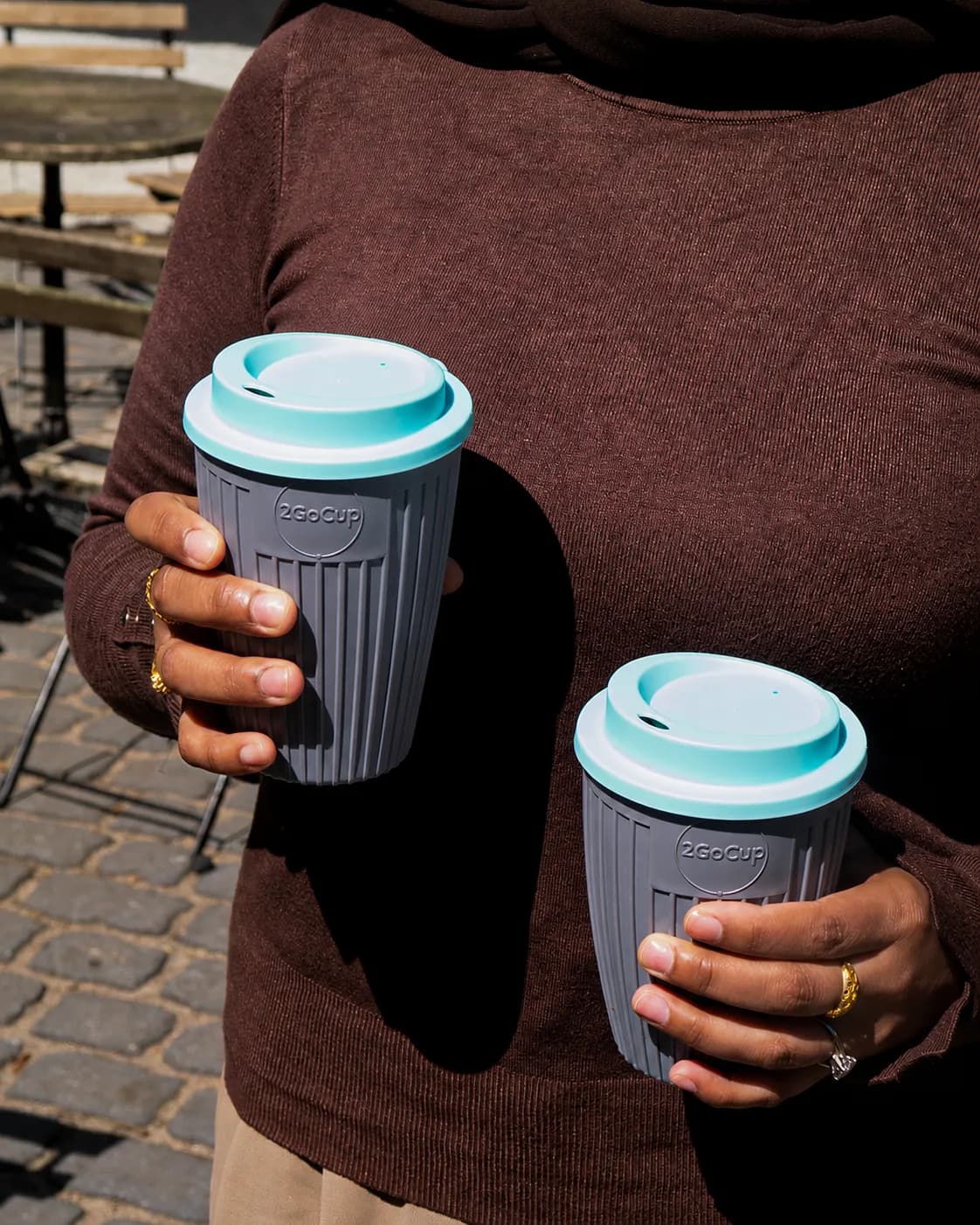 A person holds two grey reusable coffee cups with light blue lids, wearing a brown sweater and a ring on their right hand.