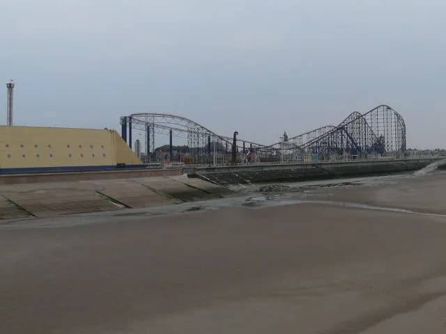 A wide shot shows a dark roller coaster against a cloudy sky, with a concrete embankment and sand in the foreground.