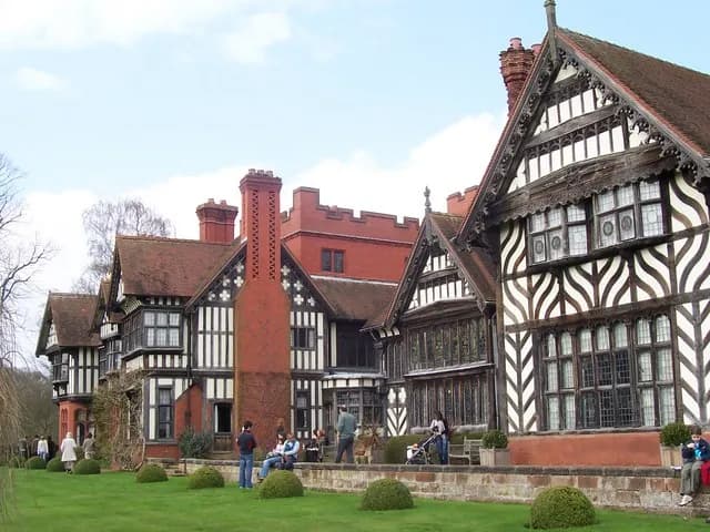 A long, black and white half-timbered mansion with red brick accents and multiple gables stands behind a green lawn with trimmed shrubs. People are walking or sitting around the building.