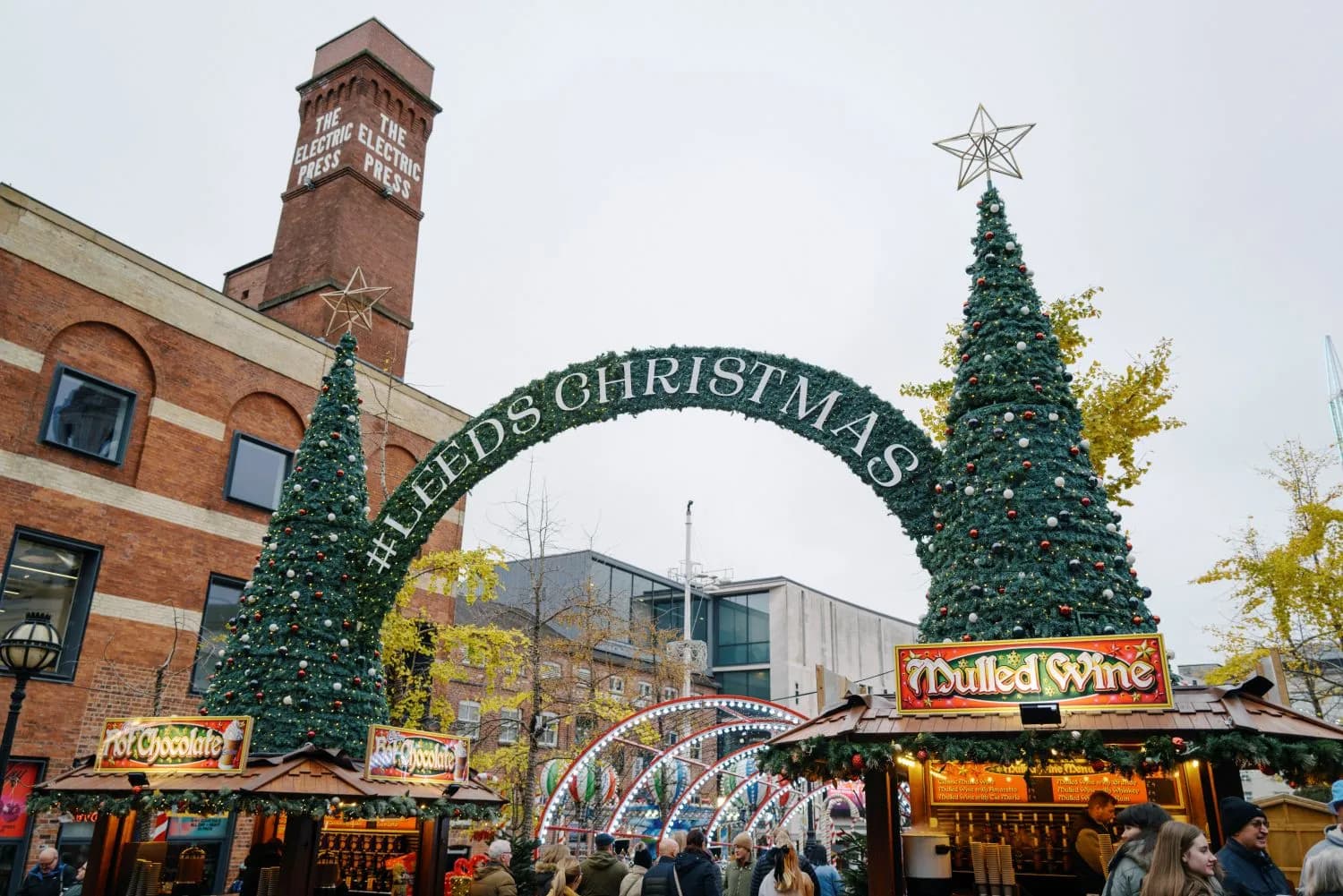 A large Christmas arch decorated with the words "#LEEDS CHRISTMAS" stands over a bustling Christmas market with stalls and people.