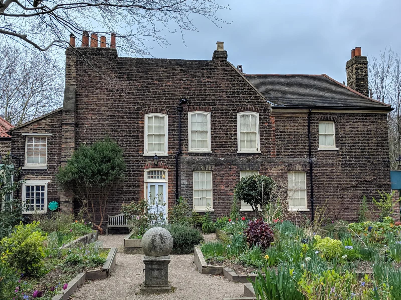 A historic brick house with white windows featuring a light blue door in the center of its facade, set against an overcast sky, with a charming garden path and various plants in the foreground.