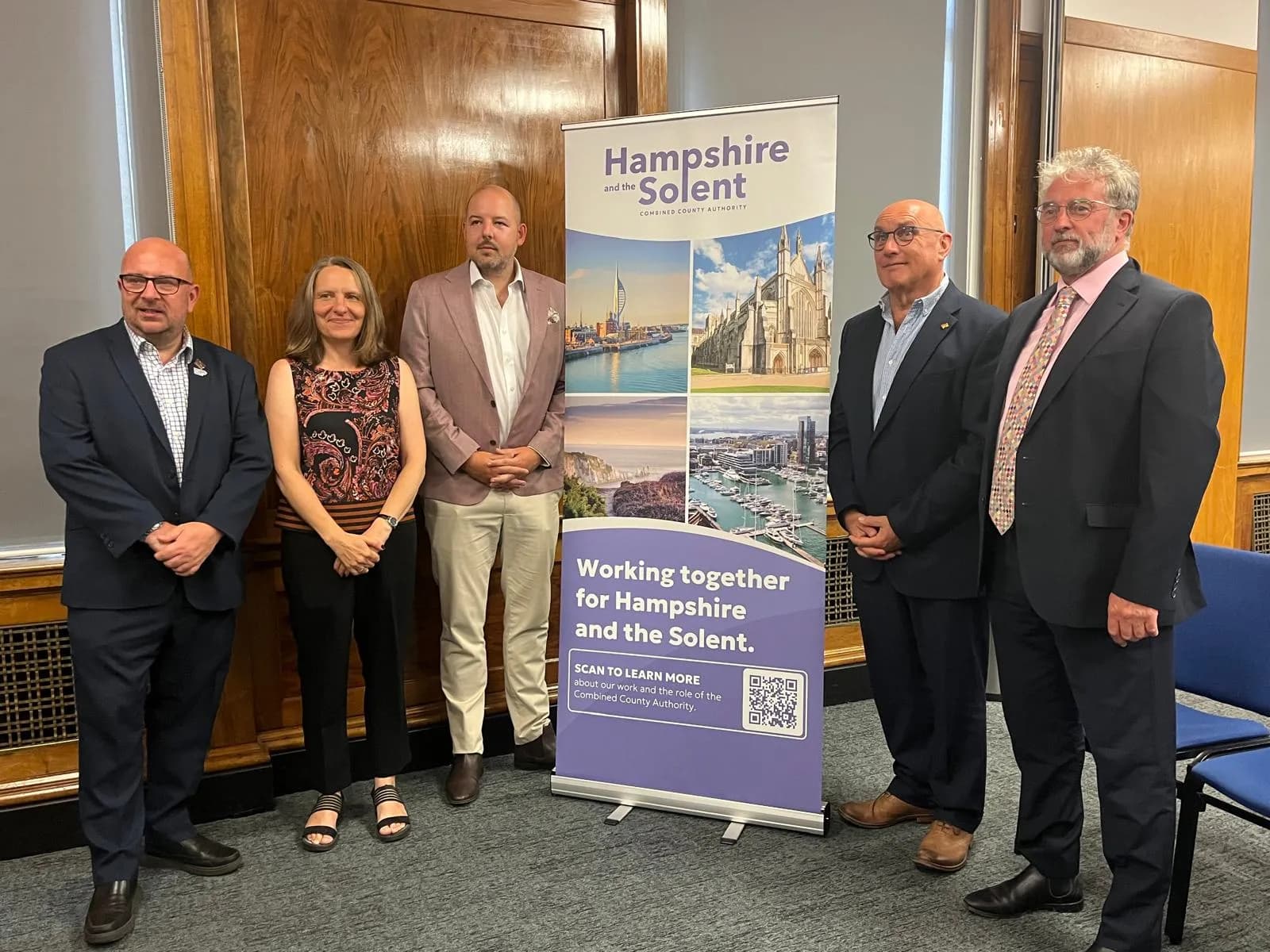 Five people, three men in suits and one woman in a patterned top, stand beside a banner promoting "Hampshire and the Solent" and discussing working together for the region.