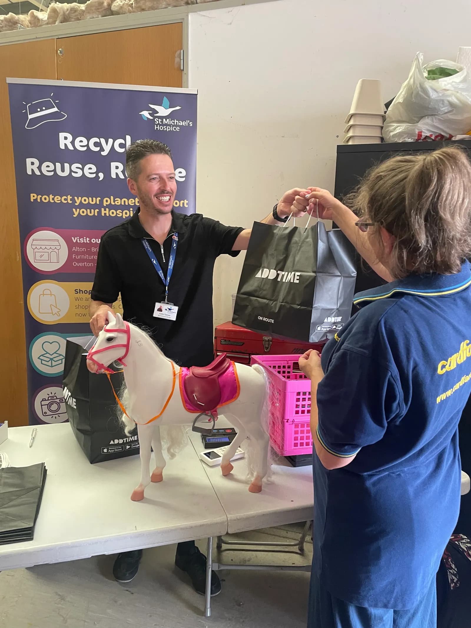 A man smiling as he hands over two shopping bags to another person, with a toy horse on the table in front of him and a banner for St Michael's Hospice behind.