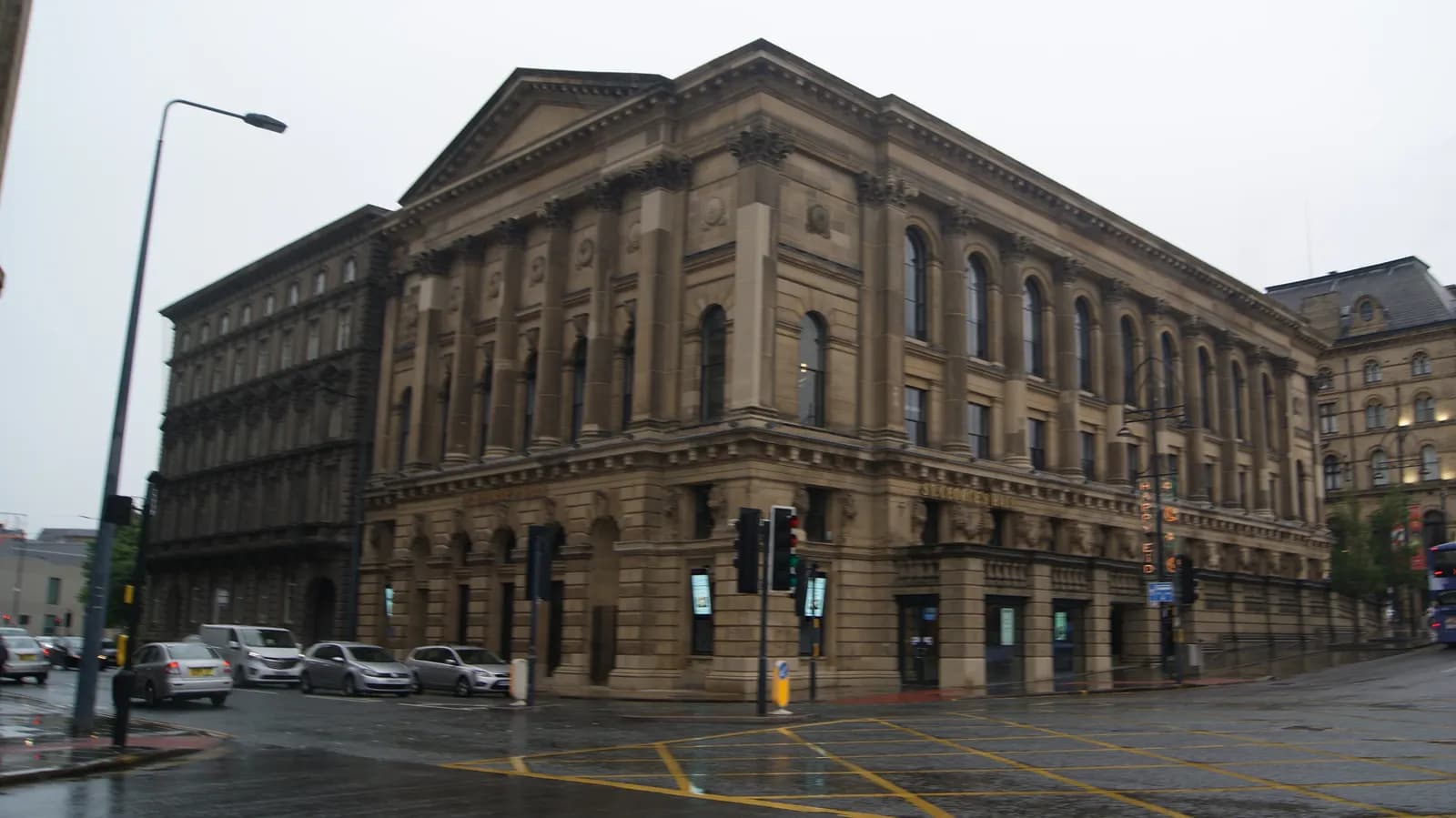 A grand, multi-story stone building with classical columns and arched windows stands at a city intersection on an overcast day, with vehicles stopped at a traffic light.