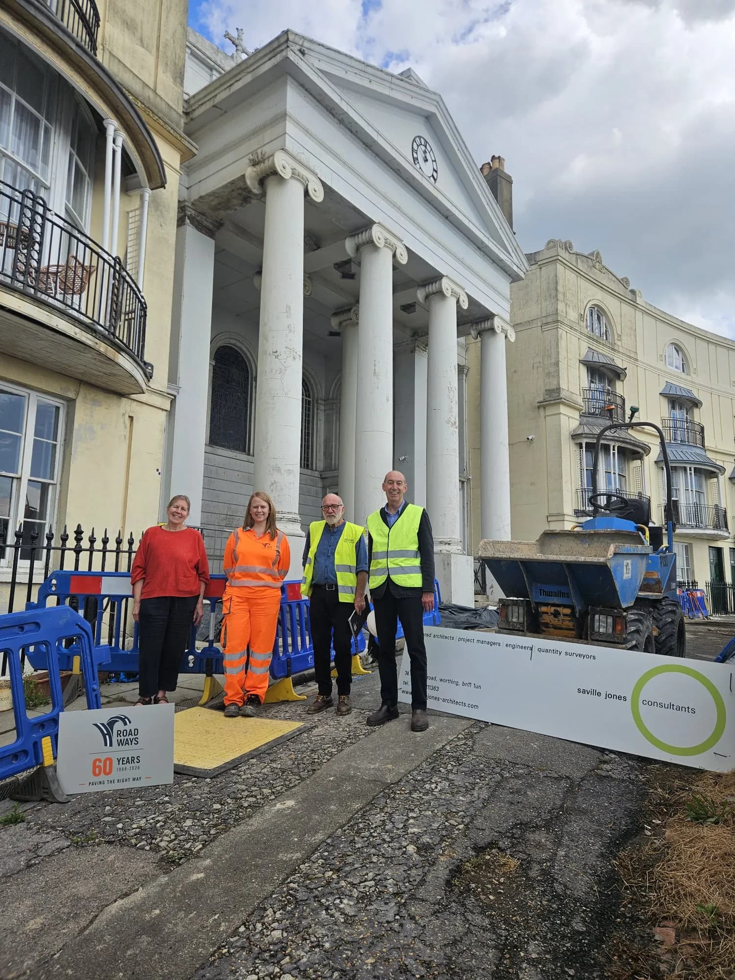 Four people, two women and two men, stand in front of a white building with large columns, with construction equipment visible to the right.