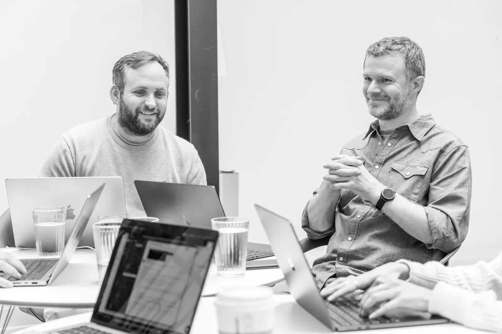 Two men are sitting across from each other at a brightly lit table with open laptops. The man on the left has a beard and is wearing a light sweater and smiling. The man on the right has a beard and is wearing a button-up shirt and a watch, with his hands clasped together, and smiling.