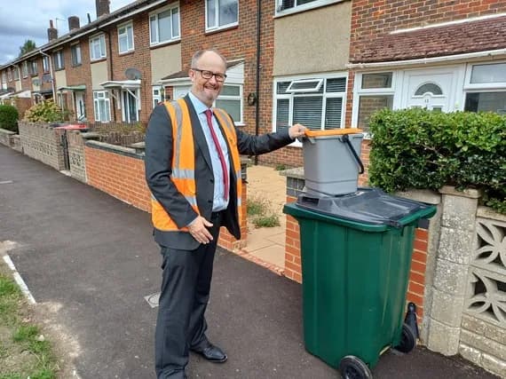 A man in a suit and an orange safety vest smiles while standing next to a green and gray recycling bin in front of a row of brick houses.