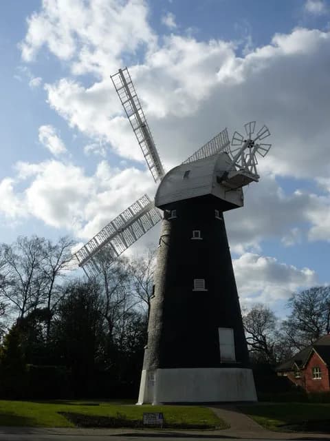 A black windmill with white rotating blades stands against a partly cloudy sky, surrounded by trees and a glimpse of a brick building.