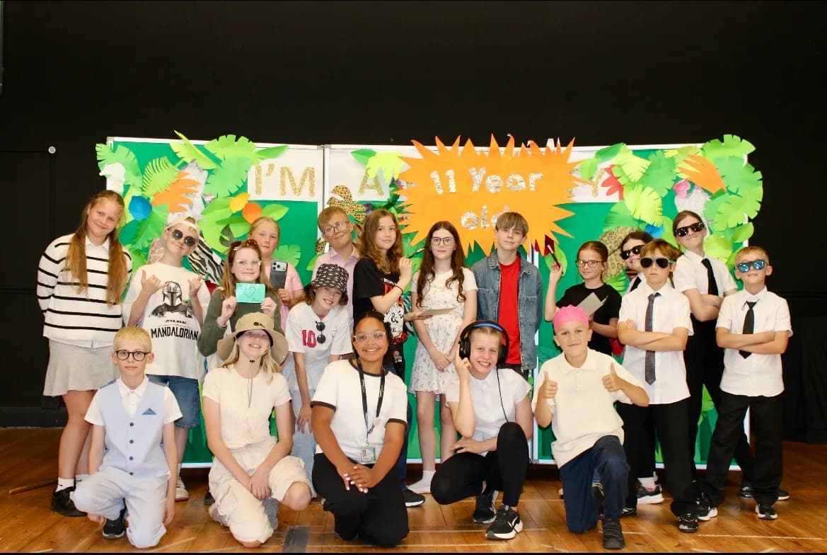 A large group of children and young teenagers stand and kneel before a green and orange tropical-themed backdrop with the words "I'M AN 11 YEAR OLD."
