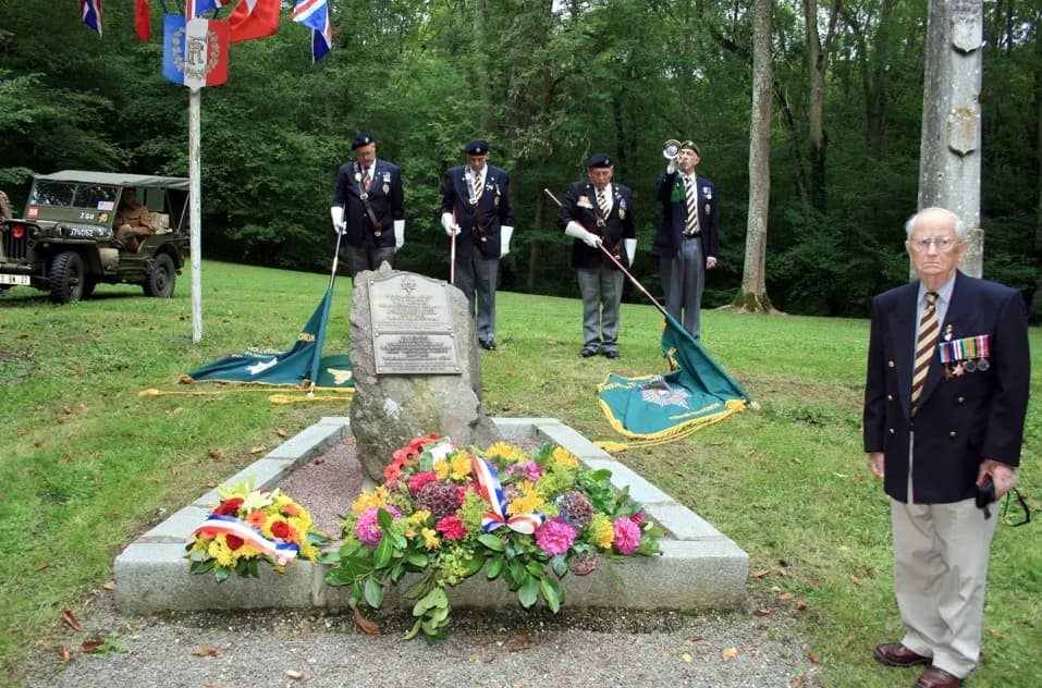 An elderly man in a suit jacket with medals stands to the right of a memorial stone decorated with flowers, while four men in uniforms stand in the background.