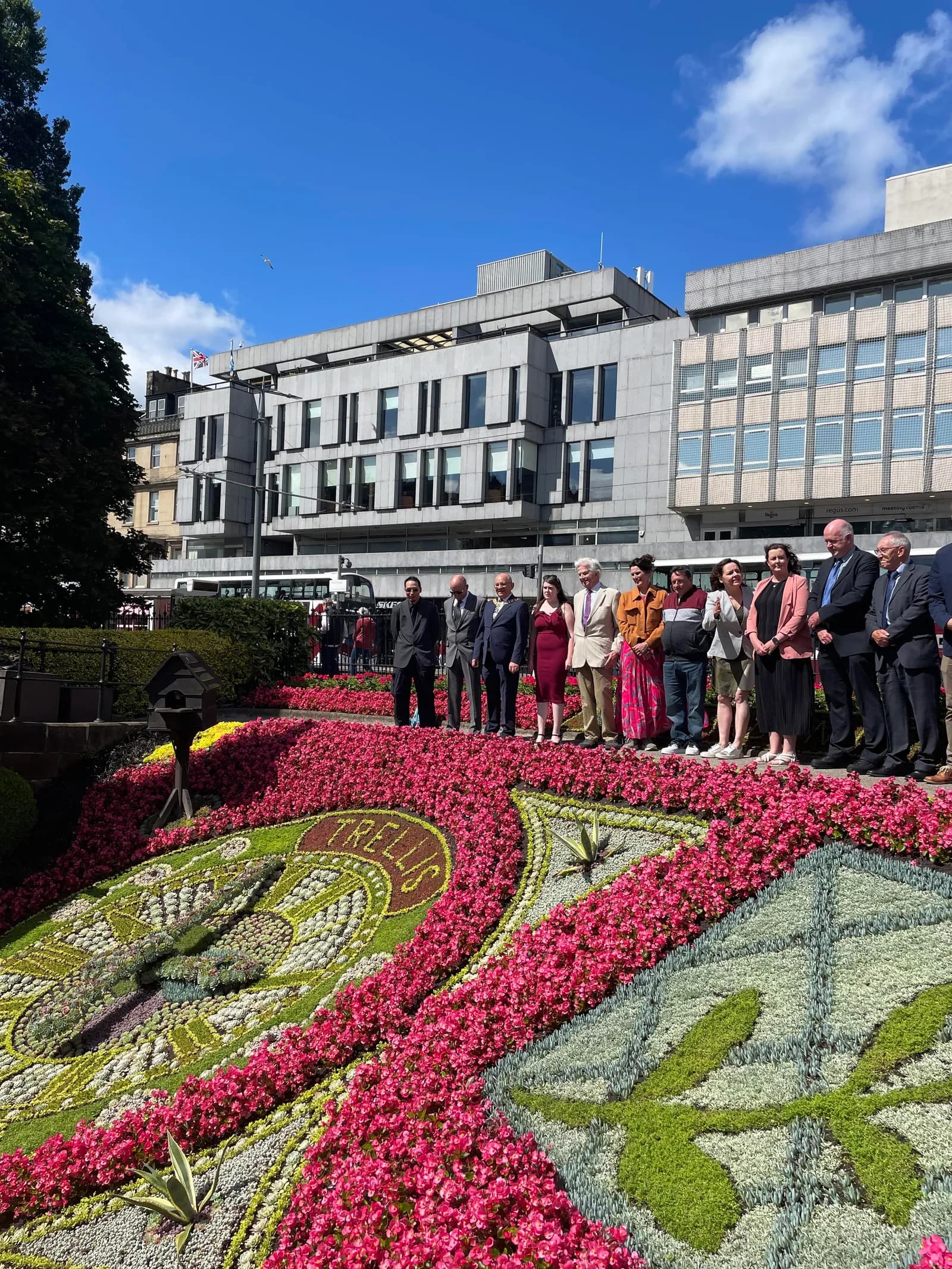 A diverse group of people stands in a line behind a large, ornate floral clock, with a modern building in the background under a blue sky.