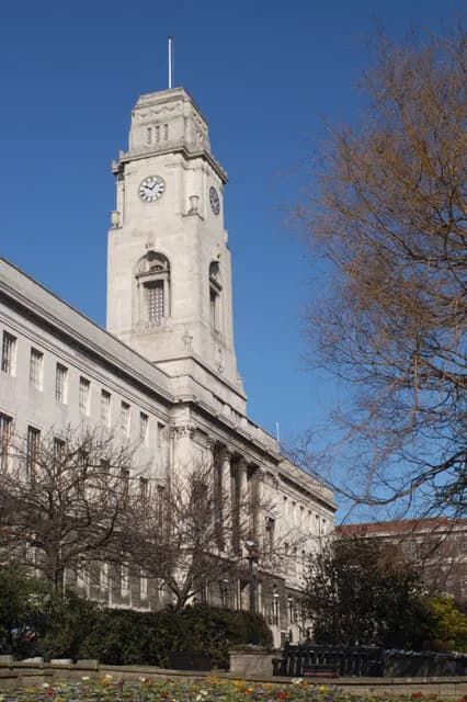 A grand municipal building with a prominent clock tower under a clear blue sky, flanked by bare trees and a colorful flower bed in the foreground.