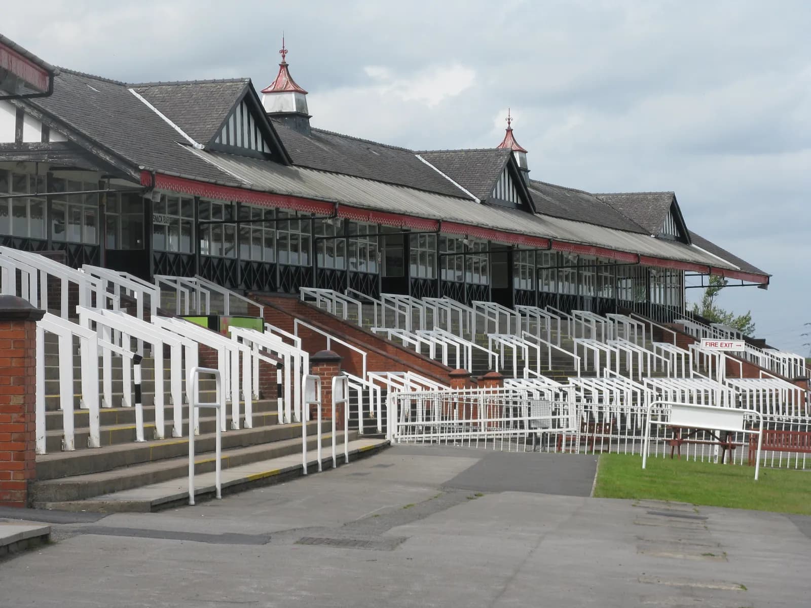 A long, single-story building with a dark roof and white-framed windows overlooks a series of tiered concrete steps with white railings and a paved ground area leading to a grassy section.