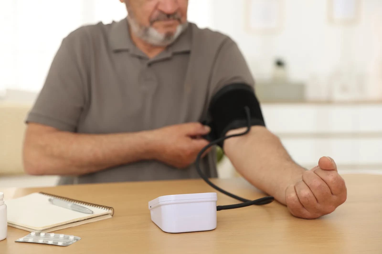 A man wearing a grey polo shirt measuring his blood pressure at home with an automatic blood pressure monitor, with a notebook and medicine blister pack on the table.