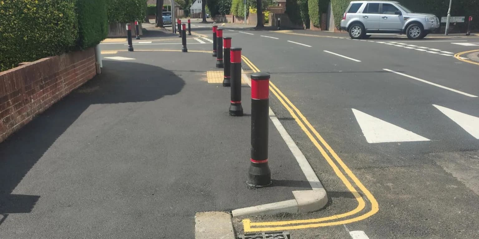 A row of black and red bollards separates a paved sidewalk from a street with double yellow lines and a white arrow marking, with a silver SUV parked in the background.