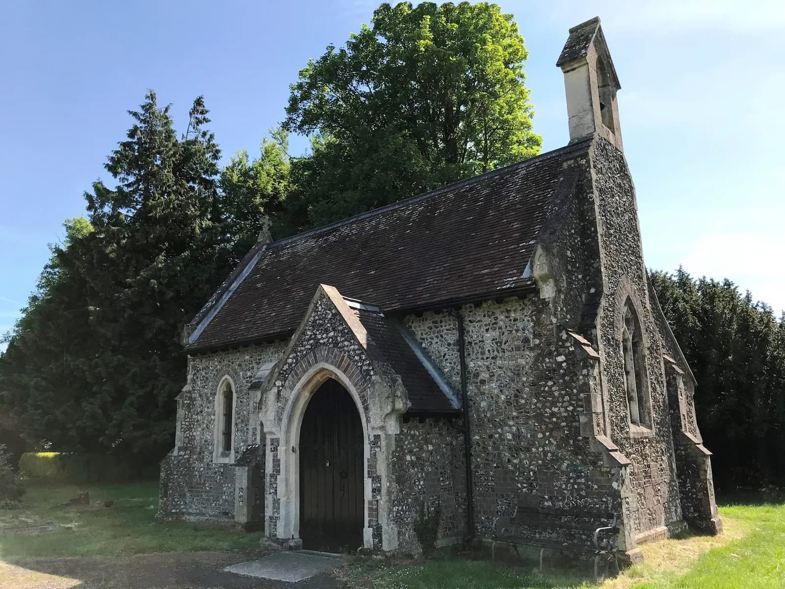 A small, old stone church with a dark roof and a prominent arched doorway stands amidst green grass and tall trees under a bright blue sky.