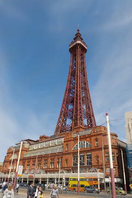 The Blackpool Tower stands tall on a sunny day, with its red iron framework contrasting against a clear blue sky, towering over the red-brick building at its base and a busy street with pedestrians and vehicles.