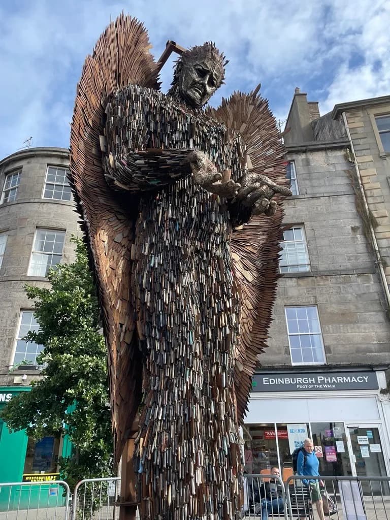 A towering angel sculpture, made from thousands of knives, stands prominently against a cloudy sky with old stone buildings and a pharmacy visible in the background.
