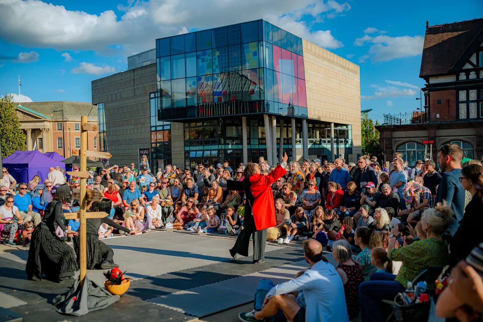 A crowd of people watches performers on a street in front of a modern glass-fronted building under a blue sky.