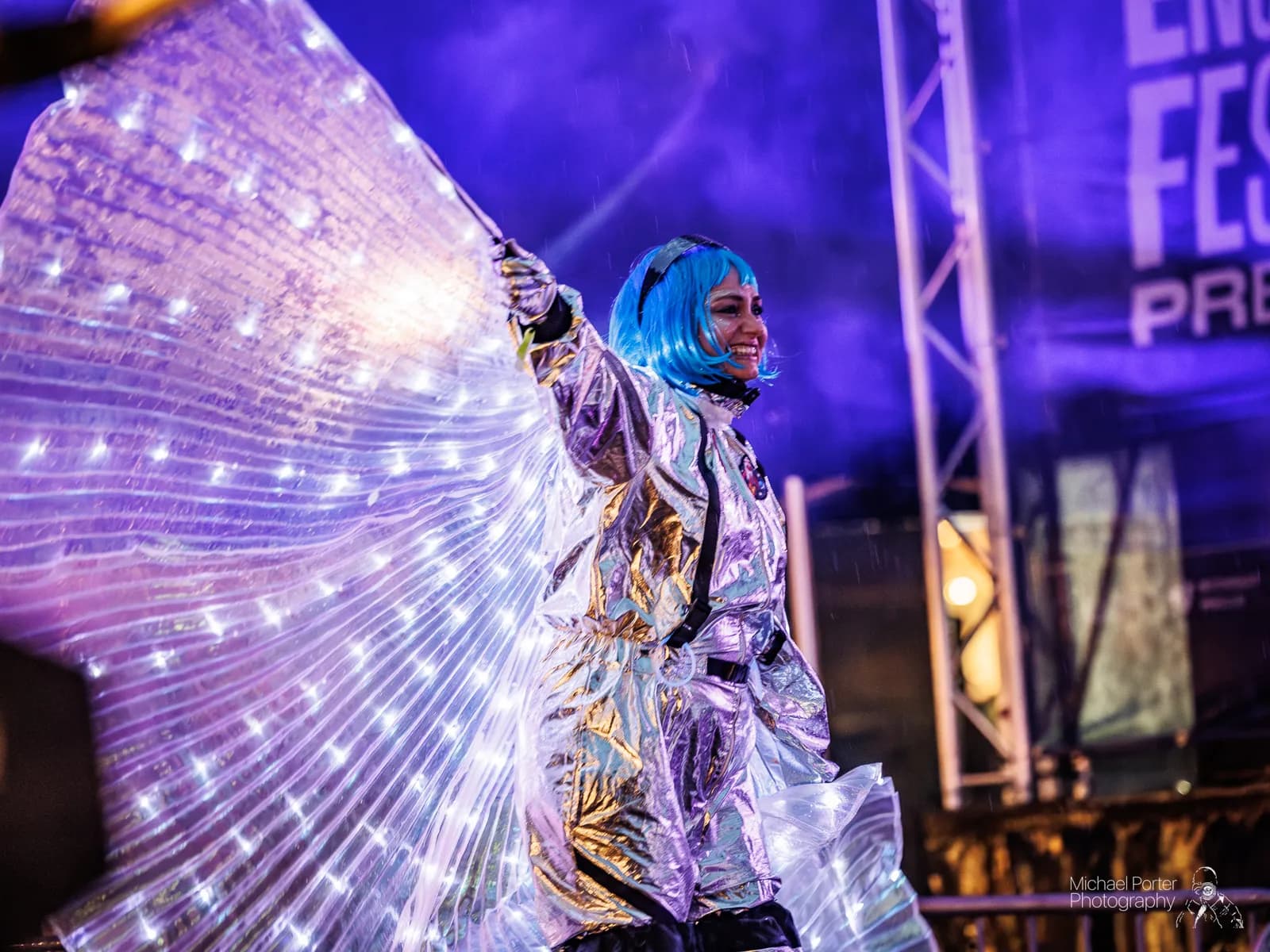 A smiling woman with blue hair and a shiny silver suit performs with a large set of illuminated wings.