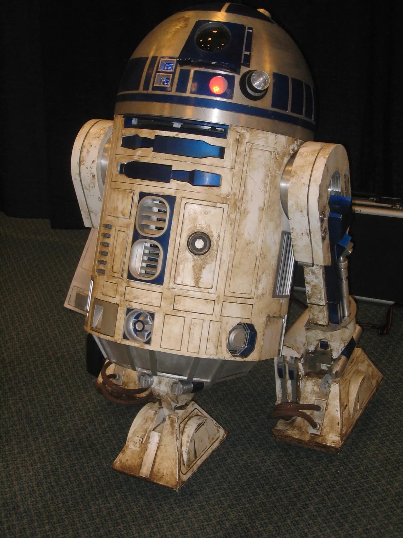 A life-sized replica of the Star Wars character R2D2, looking worn and dirty, stands on a dark carpet.