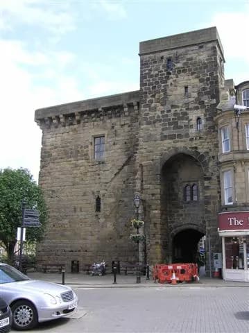 A historic stone gatehouse with a large archway over a street, partially obscured by a car and traffic barriers, stands next to a building with a red storefront.