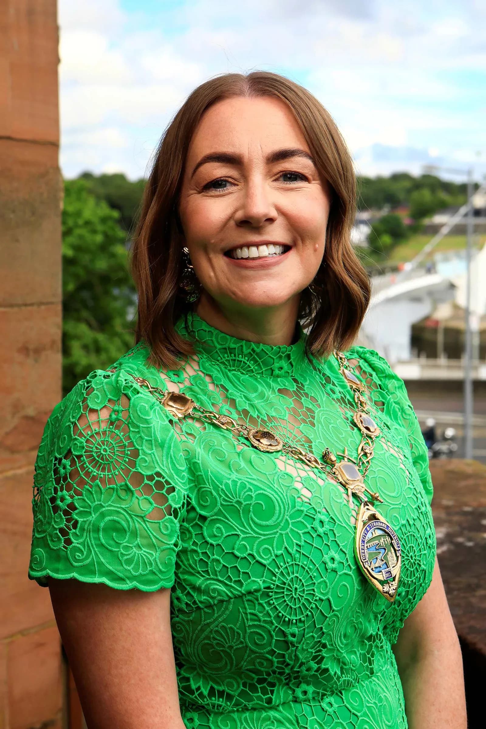 A smiling woman with brown hair and a green lace dress wears a gold chain of office with a large medal, standing outdoors with a blurred cityscape in the background.