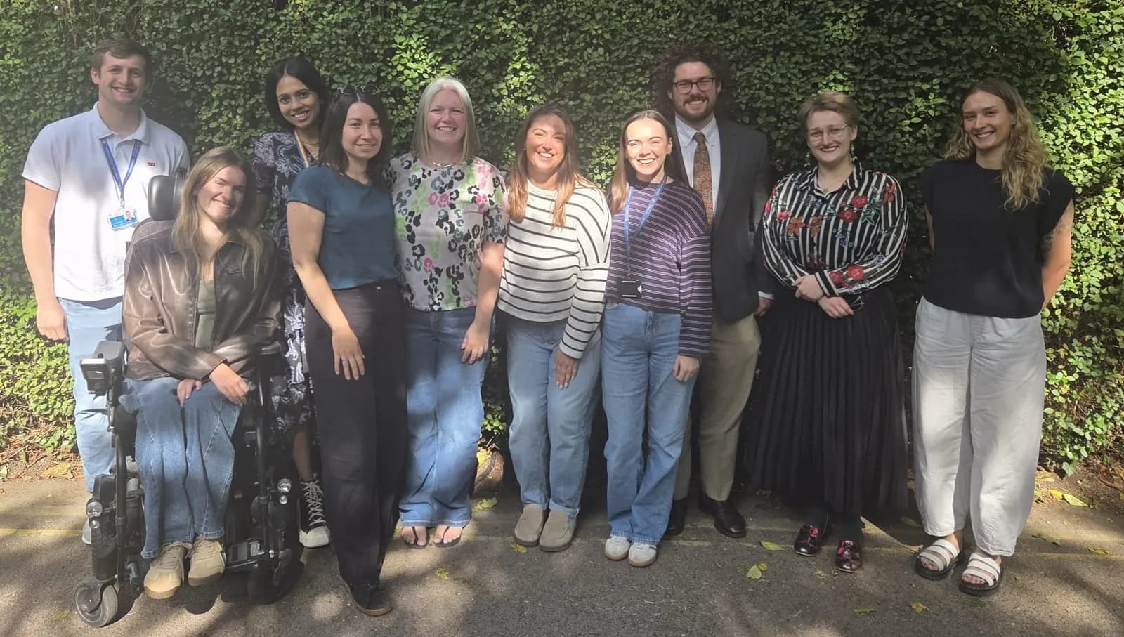 A diverse group of eleven people, some standing and one in a wheelchair, are smiling for a group photo in front of a green hedge.