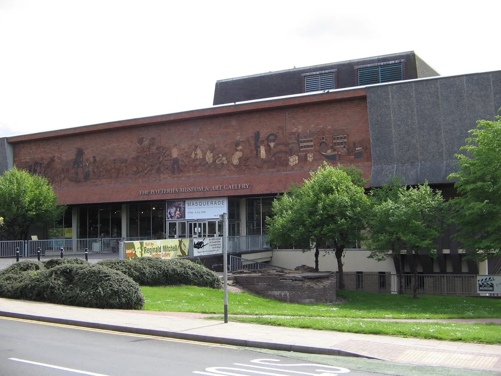 The exterior of The Potteries Museum & Art Gallery features a large brick facade with a mural depicting historical scenes, surrounded by green trees and bushes, with part of a paved street and sidewalk in the foreground.